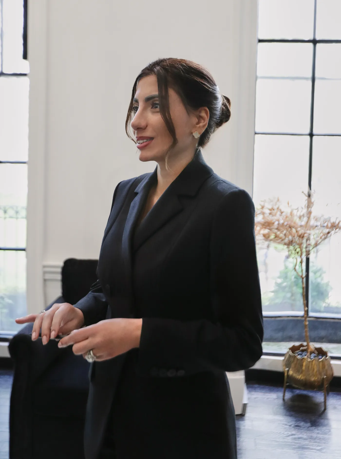 Amna Zaidi in a dark tailored suit, smiling in a bright room with tall windows